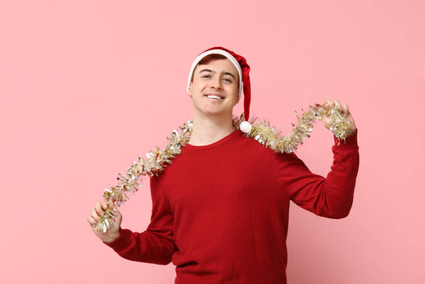 Handsome young man in warm sweater, Santa hat and tinsel on pink background - Photo, Image