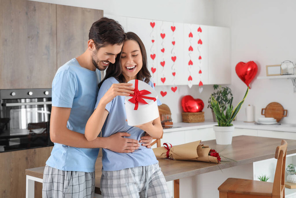 Glückliche junge Frau öffnet Geschenkbox ihres Mannes am Valentinstag in der Küche - Foto, Bild