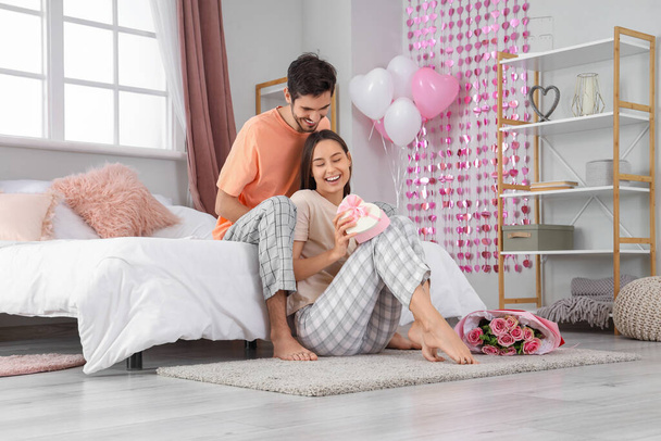 Joyeux jeune couple avec boîte cadeau dans la chambre à coucher le jour de la Saint-Valentin - Photo, image