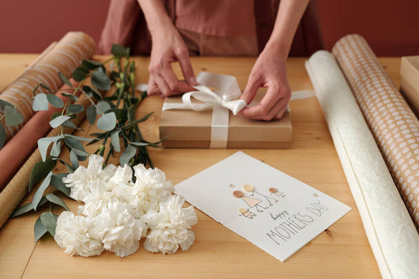 Handen van jonge onherkenbare vrouw bindt wit satijnen lint op verpakte geschenkdoos met cadeau voor haar moeder tijdens de voorbereiding voor vakantie - Foto, afbeelding