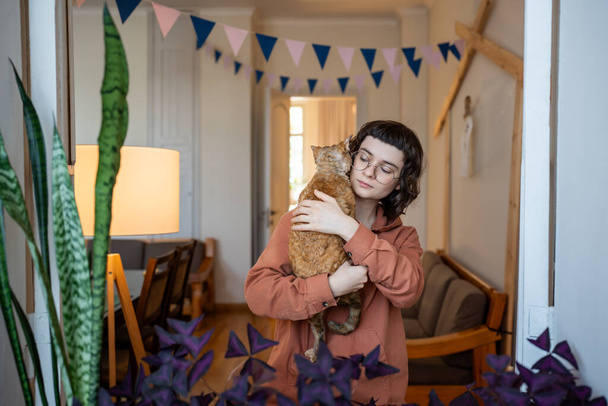 Milující teenagerka držící kočku Devon Rex v rukou, hladící ji jemně po zádech. Chlupatá zrzavá kočička sedí na dívčím rameni. Přátelství, něha, bezpodmínečná láska k domácím zvířatům, emocionální podpora zvířat - Fotografie, Obrázek