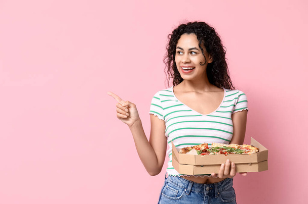 Bella giovane donna afro-americana felice con deliziosa pizza che punta a qualcosa su sfondo rosa - Foto, immagini