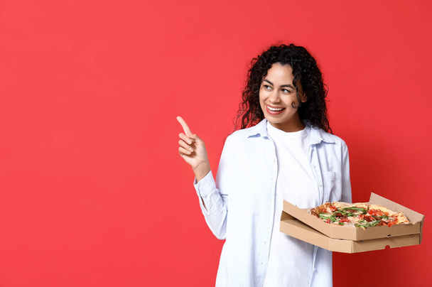 Schöne junge, glückliche Afroamerikanerin mit leckerer Pizza, die auf etwas auf rotem Hintergrund zeigt - Foto, Bild