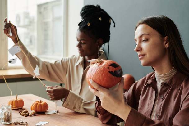 Young brunette woman looking at spooky Halloween pumpkin in her hands while sitting against female friend creating decorations - Photo, Image