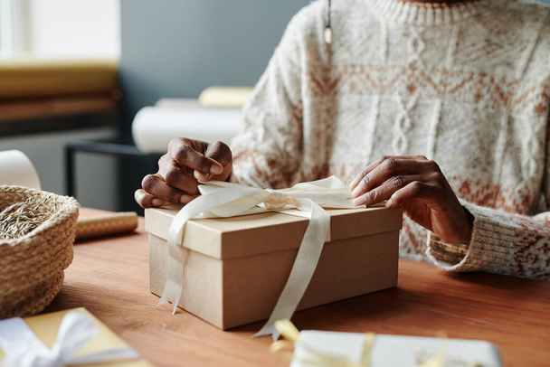 Young African American woman in sweater tying up white silk ribbon on top of giftbox cover while preparing xmas presents for her friends - Photo, Image