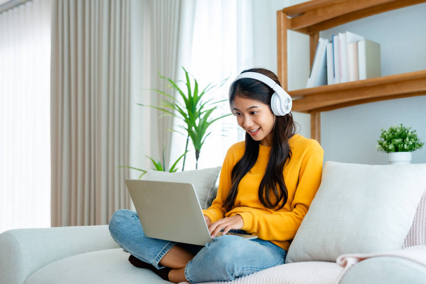 Nuori suloinen aasialainen nainen hymy yllään kuulokkeet työskentelevät olohuoneessa kotona. Onnellinen upea nainen tietokoneella kannettavan ja istuu sohvalla kotona, Videopuhelu ystävän kanssa - Valokuva, kuva