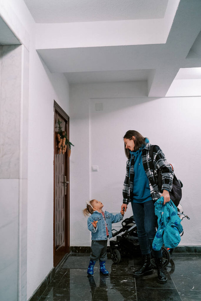 Máma se podívá na holčičku, která s ní stojí před dveřmi bytu vedle kočárku. Kvalitní fotografie - Fotografie, Obrázek