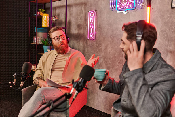 Entrevistador guapo con un atuendo elegante discutiendo preguntas con su joven invitado durante el podcast - Foto, imagen