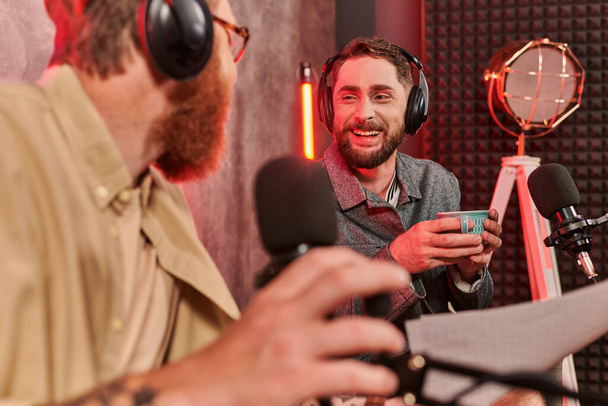 twee bebaarde knappe mannen in casual kleding met koptelefoon praten tijdens podcast, koffiekop - Foto, afbeelding