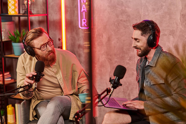 hombres guapos en atuendos casuales posando con café y portátil durante su discusión en podcast - Foto, imagen