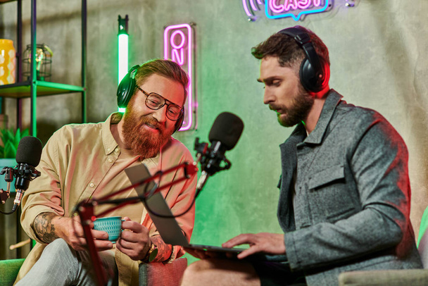 ελκυστικούς χαρούμενους άνδρες σε casual ενδυμασίες με καφέ και laptop κατά τη διάρκεια της συζήτησής τους στο podcast - Φωτογραφία, εικόνα