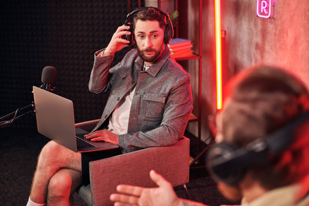 bell'uomo barbuto in abbigliamento casual accogliente seduto con computer portatile in studio accanto al suo podcaster - Foto, immagini
