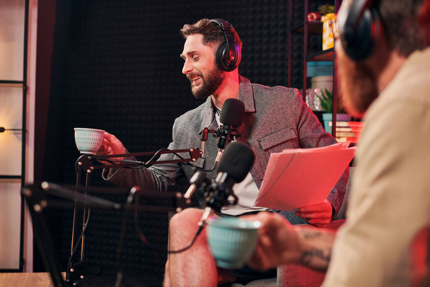 hombre barbudo alegre con taza de café en la mano mirando el papeleo al lado de su podcaster - Foto, imagen