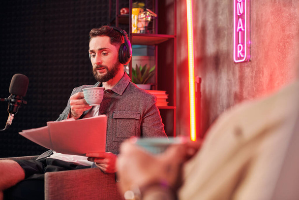hombre barbudo bien parecido con taza de café en la mano mirando el papeleo al lado de su podcaster - Foto, imagen