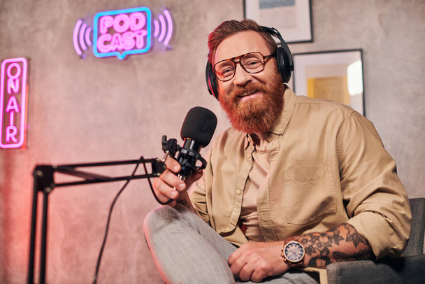homem bonito alegre com cabelo vermelho e barba com fones de ouvido olhando para a câmera durante o podcast - Foto, Imagem