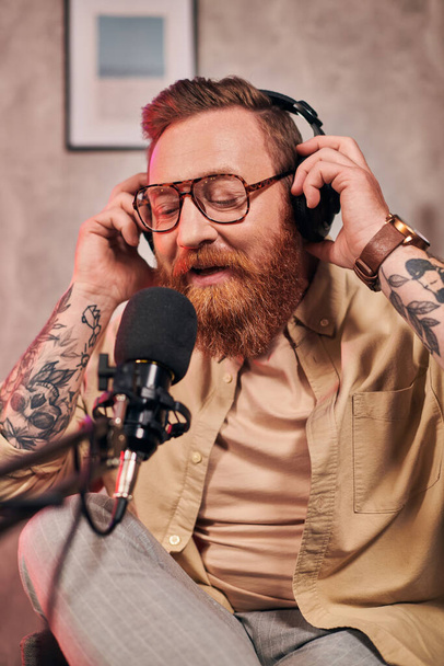 schöner Mann mit roten Haaren und Kopfhörern in eleganter Kleidung schaut während eines Podcasts in die Kamera - Foto, Bild