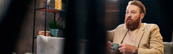 pohledný vousatý muž s rusými vlasy v elegantním oblečení sedí na pohovoru ve studiu, banner - Fotografie, Obrázek
