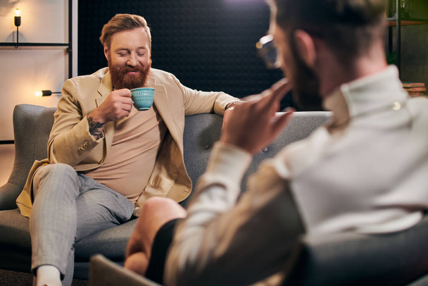 весёлый бородатый мужчина с рыжими волосами в элегантной одежде сидит рядом со своим интервьюером в студии - Фото, изображение
