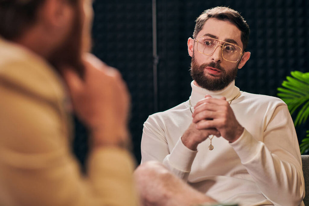 Yakışıklı, sakallı, gözlüklü, şık bir adam oturuyor ve röportajcısına bakıyor. - Fotoğraf, Görsel