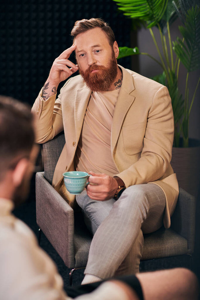 goed uitziende man met baard met koffiekopje zitten tijdens de discussie naast zijn interviewer - Foto, afbeelding