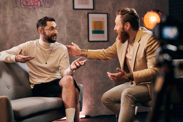 zwei attraktive bärtige Männer in eleganter, stylischer Kleidung sitzen und diskutieren Interviewfragen - Foto, Bild
