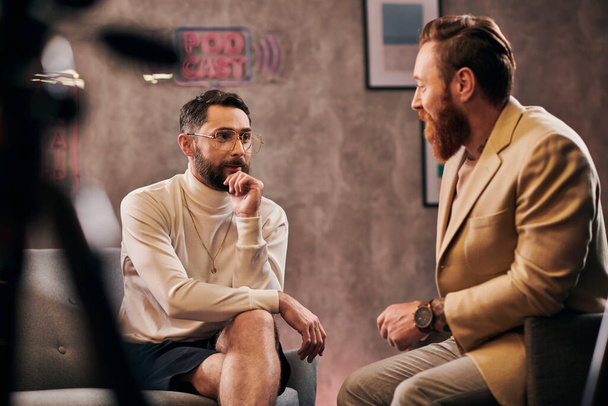 два привлекательных бородатых мужчины в элегантных стильных одеждах сидя и обсуждая вопросы интервью - Фото, изображение