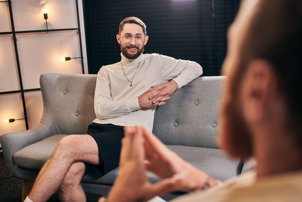 cheerful bearded men in casual outfit smiling and looking at his interviewer while sitting in studio - Photo, Image