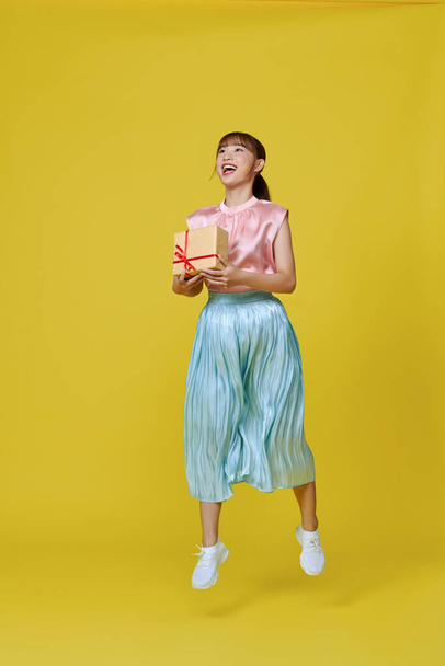 Szczęśliwy ładna młoda kobieta skacząc i trzymając opakowane pudełko z uśmiechem odizolowany na żółtym tle - Zdjęcie, obraz