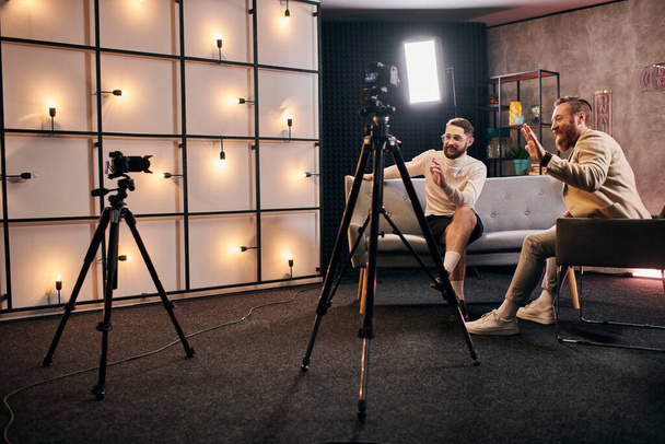 homens elegantes bonitos com barbas em trajes chiques acenando para a câmera durante a entrevista em estúdio - Foto, Imagem
