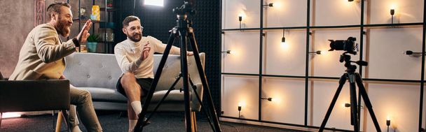 gutaussehende elegante Männer mit Bärten in schicken Anzügen winken während des Interviews im Studio in die Kamera, Banner - Foto, Bild