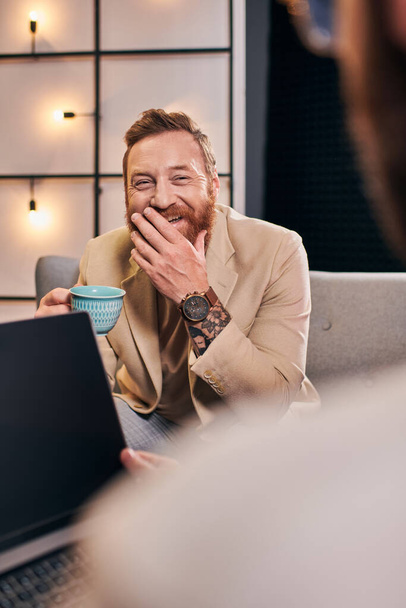 δύο χαρούμενα ελκυστική άνδρες σε κομψά ρούχα με καφέ και laptop συζητώντας ερωτήσεις συνέντευξη - Φωτογραφία, εικόνα