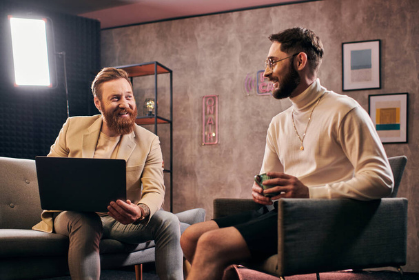 dwóch radosnych atrakcyjnych mężczyzn w eleganckich strojach z kawą i laptopem omawiających pytania o wywiad - Zdjęcie, obraz