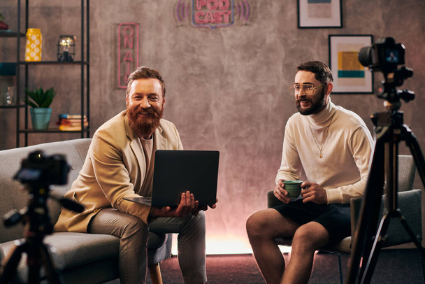 veselý pohledný muži v módní oblečení s kávou a notebookem s úsměvem během rozhovoru - Fotografie, Obrázek
