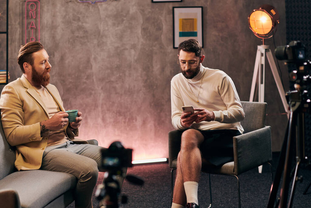 красивые элегантные мужчины в шикарных одеждах с бородами, разговаривающие во время интервью, с кофе и телефоном - Фото, изображение