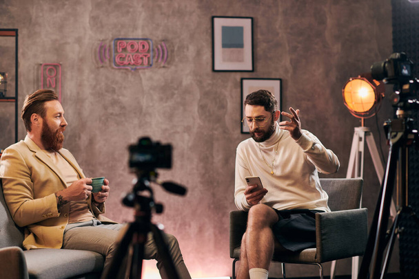 εμφανίσιμοι κομψοί άνδρες με κομψές ενδυμασίες με γένια που μιλάνε κατά τη διάρκεια της συνέντευξης, με καφέ και τηλέφωνο - Φωτογραφία, εικόνα
