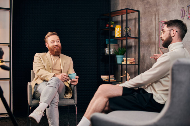 vrolijke knappe mannen in elegante kleding met koffie bespreken vragen tijdens interview - Foto, afbeelding