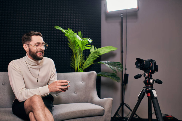 radosny brodaty mężczyzna w eleganckim stroju z okularami trzymającymi filiżankę kawy podczas wywiadu w studio - Zdjęcie, obraz