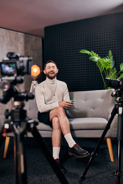 веселый бородатый мужчина в элегантной одежде с очками, держа чашку кофе во время интервью в студии - Фото, изображение