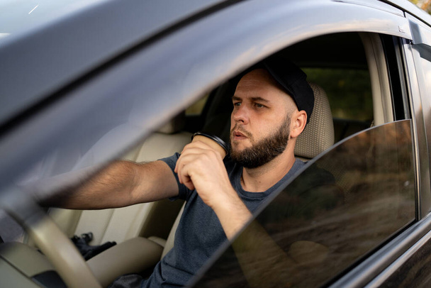 Bearded man in cap or male driver driving car and drinking takeaway coffee. lifestyle and people concept. Copy space - Photo, Image