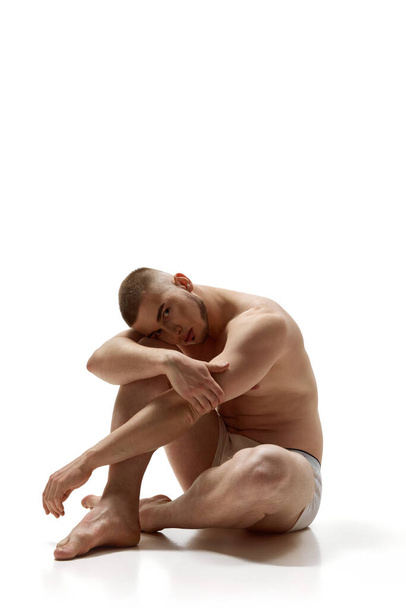 Νεαρός άνδρας, με αθλητική άνθρωπος με τέλειο σώμα κάθεται στο πάτωμα με το κεφάλι του στην αγκαλιά του σε λευκό φόντο στούντιο. Έννοια της mens υγεία, την ομορφιά, το σώμα και την περιποίηση του δέρματος, γυμναστήριο. Τέχνη σώματος - Φωτογραφία, εικόνα