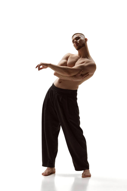 Embodying peak fitness. Athletic shirtless man defines strength and grace in black trousers against white studio background. Concept of mens health, beauty, body and skin care, fitness. Body art - Photo, image