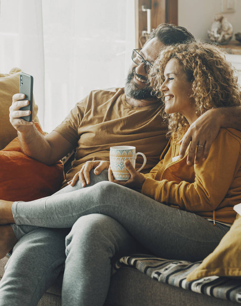 Couple of young adult man and woman enjoy time together sitting on the sofa in indoor leisure activity.. Real life people at home. Male and female relax on the couch reading a book and drinking a tea - Valokuva, kuva