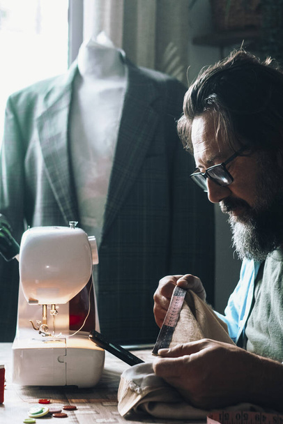 Man at home using sewing machine to tailor and modify garments. Blazer in background. Tailoring and tailor at work at the workshop. Concept of people and alternative job. Professional tailor - Photo, Image