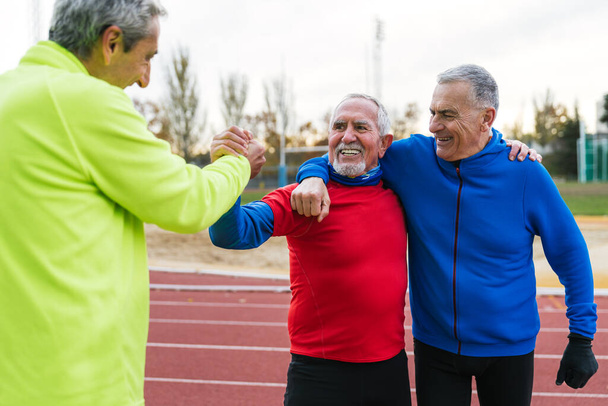 Tres alegres hombres mayores en ropa deportiva celebran la compañía en una pista de atletismo, abrazando y estrechando las manos después de un trote. - Foto, Imagen