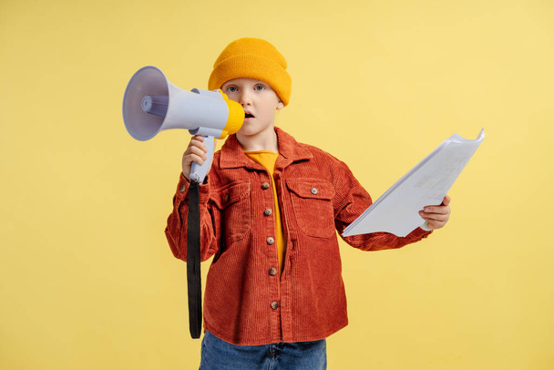 Porträt eines niedlichen kleinen Mädchens mit gelbem Hut, lässiger brauner Jacke, die Papier und Lautsprecher in der Hand hält, redet und die Kamera isoliert auf gelbem Hintergrund betrachtet. Kommunikationskonzept, Protest - Foto, Bild