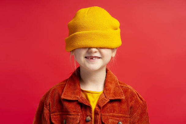 Portrét roztomilé holčičky se žlutým kloboukem a neformálním oblečením, poflakující se kolem, usmívající se osamoceně na červeném pozadí. Dětský koncept - Fotografie, Obrázek