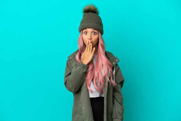 手で口を覆う青い背景に隔離された防水コートを着用したピンクの髪の若い女性 - 写真・画像