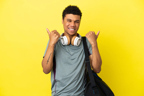 Joven deporte hombre afroamericano con bolsa de deporte aislado sobre fondo amarillo con los pulgares hacia arriba gesto y sonriendo - Foto, Imagen