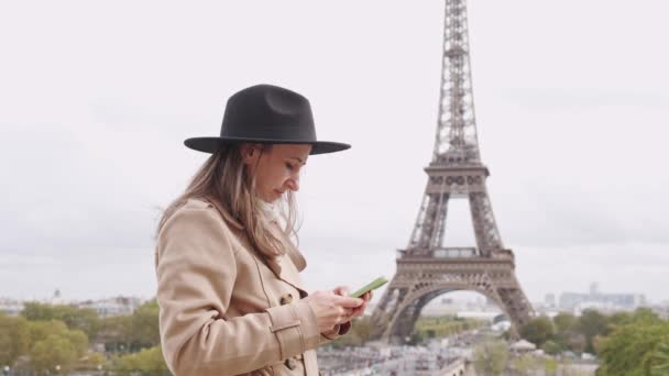Πλάνα νεαρής γυναίκας που προειδοποιεί με μαύρο καπέλο και παλτό κοντά στον Πύργο του Άιφελ χρησιμοποιώντας το smartphone της. - Πλάνα, βίντεο