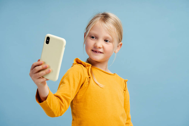 Portrait of beautiful smiling little girl, influencer holding mobile phone, taking selfie, recording video, social media, posing isolated on blue background. Technology concept - Photo, Image